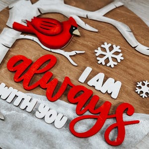 K&ouml;nnte beinhalten: Ein rundes Holzschild mit einem roten Kardinal, der auf einem wei&szlig;en Ast sitzt. Das Schild hat einen wei&szlig;en, schneebedeckten Hintergrund mit den Worten "Ich bin immer bei dir" in roten und wei&szlig;en Buchstaben.