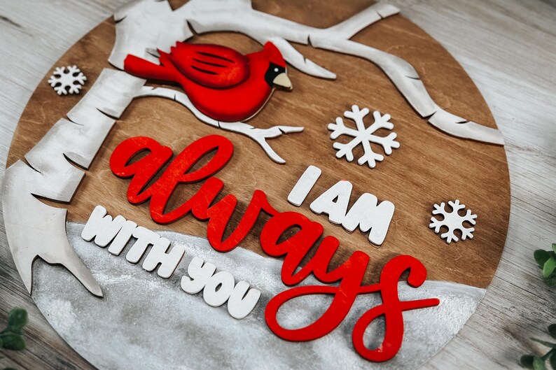 K&ouml;nnte beinhalten: Ein rundes Holzschild mit einem roten Kardinal, der auf einem wei&szlig;en Ast sitzt. Das Schild hat einen wei&szlig;en, schneebedeckten Hintergrund mit den Worten "Ich bin immer bei dir" in roten und wei&szlig;en Buchstaben.