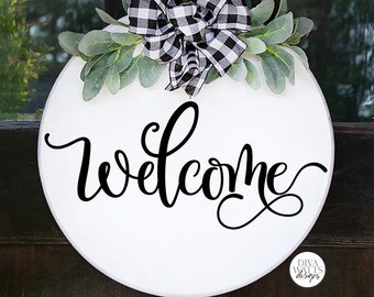 May include: White round wooden sign with a black and white checkered bow and greenery. The sign says "Welcome" in black cursive lettering.