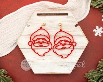 May include: Red Santa Claus face earrings with gold-colored ear wires. The earrings are displayed on a white and wood-toned hexagonal display. The background is a red surface with white snowflakes and greenery.