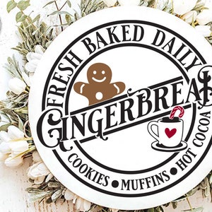 May include: A white round sign with black text that reads "Fresh Baked Daily Gingerbread Cookies Muffins Hot Cocoa". A gingerbread man cookie and a cup of hot cocoa with a candy cane are illustrated on the sign.