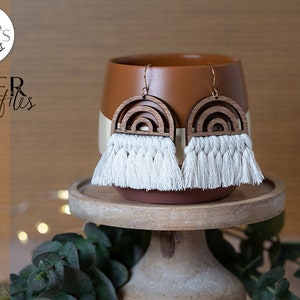 May include: A pair of wooden rainbow earrings with white tassel fringe. The earrings are displayed on a brown ceramic pedestal.