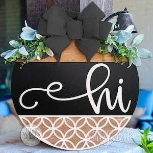 Könnte beinhalten: Runder Willkommensgruß aus Holz mit schwarzer Mitte und dem Wort "hi" in weißer Schrift. Die Oberseite ist holzfarben, mit einer schwarzen Schleife und Grün. Der Boden hat ein dekoratives geometrisches Muster.