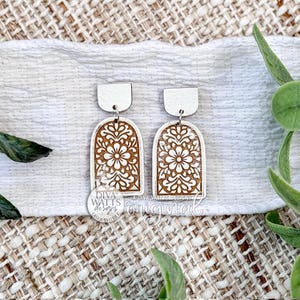 May include: A pair of wooden earrings with a floral design. The earrings have a white top and a brown, arched bottom with a detailed floral pattern. The earrings are displayed on a white cloth with green leaves in the background.