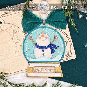 May include: A wooden snowman ornament with a blue scarf and a snow globe design. The ornament is unfinished and ready to be painted. The text "Let it Snow!" is written on the ornament.