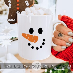 Peut inclure: Une tasse en c&eacute;ramique blanche avec un motif de visage de bonhomme de neige. Le bonhomme de neige a des yeux noirs, un nez de carotte orange et un sourire noir. La tasse est remplie de guimauves et d'un biscuit au pain d'&eacute;pices brun.