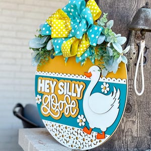 May include: A round wooden door hanger with a yellow, teal, and white design. It features a white goose with orange feet and the text "Hey Silly Goose" in yellow and teal lettering.