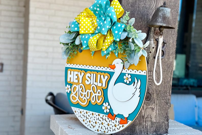 May include: A round wooden door hanger with a yellow, teal, and white design. It features a white goose with orange feet and the text "Hey Silly Goose" in yellow and teal lettering.