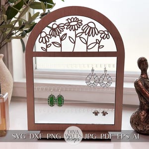 Peut inclure: Un présentoir à boucles d'oreilles en bois avec un motif floral. Le présentoir a trois étagères pour les boucles d'oreilles et une façade en acrylique transparente. Le présentoir est posé sur une surface blanche.