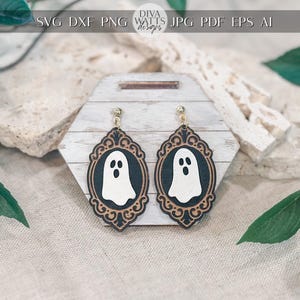 Op de afbeelding: Een paar Halloween-oorbellen. Elke oorbel heeft een wit spookontwerp in een zwart ovaal frame met goudkleurige details. De oorbellen worden op een houten standaard getoond. Het Diva Watts Designs-logo is zichtbaar.