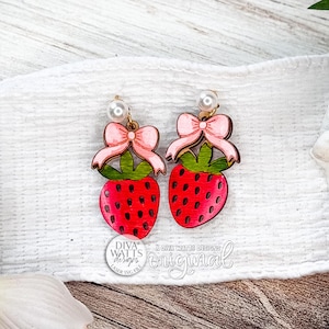 May include: A pair of wooden strawberry earrings. Each earring features a red strawberry with green leaves, a pink bow, and a pearl accent. The earrings are set against a white background.