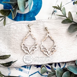 May include: Two light wood-toned earrings with an openwork, ornate design. The earrings hang from small silver-coloured posts. The image includes the text "DIVA WATTS DESIGNS" and "original". The earrings rest on a white cloth with blue floral patterns and greenery.