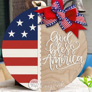 May include: A round wooden sign featuring the American flag design on one half and the phrase "God Bless America" on the other. The sign is adorned with a red, white, and blue bow. The sign is a patriotic decoration.
