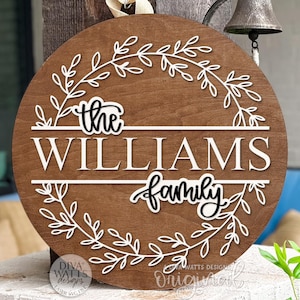 Puede incluir: Un letrero redondo de madera con el nombre de la familia, con un acabado marrón oscuro. El letrero presenta las palabras "the WILLIAMS family" en blanco, con un diseño de corona floral blanca. Adecuado para uso en interiores o exteriores.
