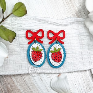 May include: Wooden earrings featuring a red bow and a strawberry design. The earrings have a blue scalloped border and a white background with a dotted pattern. The strawberries are red with green tops.