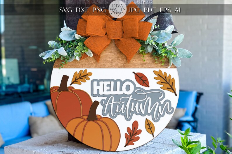 May include: A round wooden fall-themed door hanger with a white and brown design. It features orange pumpkins, fall leaves, and the words "Hello Autumn". The top has an orange bow and greenery.
