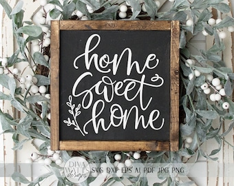 以下が含まれることがあります： 黒い背景に白い文字で「home sweet home」と書かれた木製の看板。看板は白いベリーと緑の葉で囲まれています。