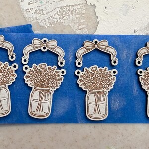 May include: Four wooden laser-cut ornaments. Each ornament features a mason jar filled with flowers, topped with a bow and a ribbon. The ornaments are light brown and are on a blue surface.