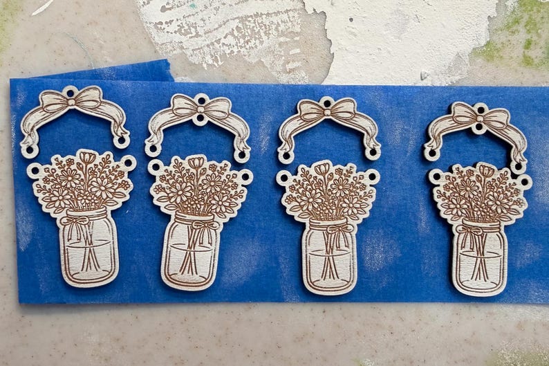 May include: Four wooden laser-cut ornaments. Each ornament features a mason jar filled with flowers, topped with a bow and a ribbon. The ornaments are light brown and are on a blue surface.