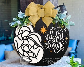 May include: A black wooden Christmas door hanger with a white cutout of the Holy Family. The words "O Night Divine" are written in white script. The hanger is decorated with a gold bow, greenery, and a small bell.