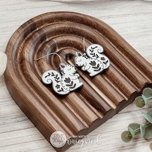 Pode incluir: Um par de brincos em forma de esquilo branco com detalhes de linhas florais pretas e ganchos de metal prateado. Os brincos estão expostos numa bandeja de madeira castanha escura em forma de arco com uma superfície estriada e um logótipo.