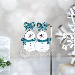 Puede incluir: Pendientes de muñeco de nieve con lazos y bufandas de purpurina color turquesa. Los muñecos de nieve blancos tienen ojos y bocas de botón negro y una pequeña nariz roja. El texto "DIVA WATTS DESIGNS" es visible.