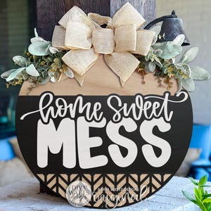 May include: A round wooden sign with the words "home sweet MESS" in white script and block letters. The sign is black and light brown, with a decorative bow and greenery at the top. A small bell is attached to the top right.