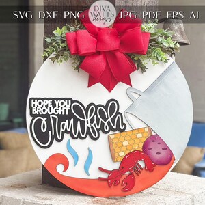 May include: A white round sign with the text "Hope you brought crawfish" in black lettering. The sign is decorated with a red bow, a white pitcher, a lobster, corn, and a purple potato.