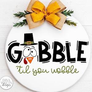 May include: A white round wooden sign with a turkey face and the text "Gobble til you wobble" in black and green lettering. A yellow and brown bow is at the top of the sign.