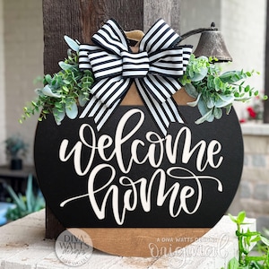 May include: A round, black wooden sign with the words "welcome home" in white script. The sign is decorated with a black and white striped bow, greenery, and a small bell. The sign is displayed outdoors.