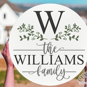 Puede incluir: Un letrero redondo blanco con la letra "W" en negro, rodeado de ramas verdes con hojas. El texto "the WILLIAMS family" está escrito en negro y gris debajo de la letra. El letrero está sostenido por las manos de una persona.