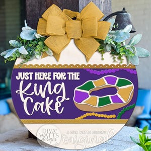 May include: A round wooden sign with a purple background and the text "JUST HERE FOR THE KING CAKE." It features a king cake graphic and is decorated with a gold bow, greenery, and a bell. The sign is a festive decoration.
