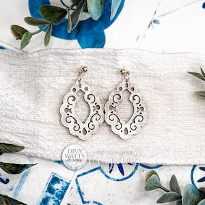 Peut inclure: Une paire de boucles d'oreilles ajourées blanches avec un motif filigrané. Les boucles d'oreilles ont une forme de goutte d'eau avec des détails de volutes complexes et de petits clous dorés. Le fond comprend de la verdure et un tissu blanc.