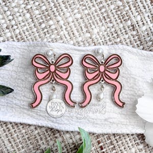 Puede incluir: Un par de pendientes de lazo rosa con contornos marrones y detalles de perlas colgantes. Los pendientes se exhiben sobre una tela blanca con detalles florales y verdes. Los pendientes tienen un perno de perla y un pequeño dije.