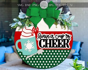 Pode incluir: Uma placa redonda de madeira com um laço verde, vegetação e o texto "have a cup of CHEER". A placa apresenta uma seção listrada em vermelho e branco, uma seção de bolinhas verdes e uma caneca azul-turquesa com um design de floco de neve.