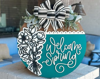 May include: A teal round wooden door hanger with a white scalloped edge. The door hanger has a white cursive inscription that says "Welcome Spring" and a black and white floral design on the left side. A black and white striped bow is attached to the top of the door hanger.