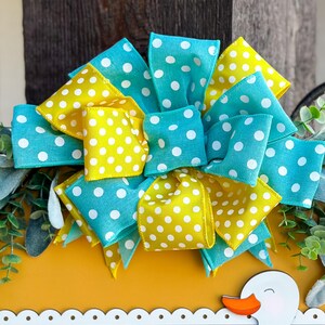 May include: A yellow and blue polka dot bow with a white background. The bow is made of ribbon and is tied in a knot. The bow is sitting on top of a yellow surface.