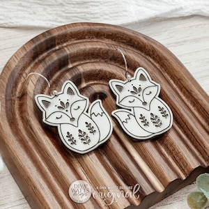 Könnte beinhalten: Ein Paar weißer, fuchs-förmiger Ohrhänger mit schwarzen, gravierten floralen und geometrischen Details. Jeder Ohrring hat einen silberfarbenen Metallhaken und zeigt einen schlafenden Fuchs mit geschlossenen Augen und buschigem Schwanz.
