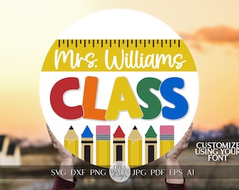 Puede incluir: Un cartel redondo blanco con una regla amarilla en la parte superior y el texto "Mrs. Williams CLASS" en letras de colores. El cartel está decorado con una fila de lápices amarillos con gomas de borrar rojas, azules y rosas. El cartel está siendo sostenido por las manos de una persona.