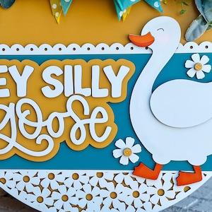 May include: A round wooden sign with a teal background and a white goose cutout. The sign says "Hey Silly Goose" in white letters on a yellow background.