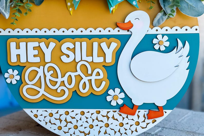 May include: A round wooden sign with a teal background and a white goose cutout. The sign says "Hey Silly Goose" in white letters on a yellow background.