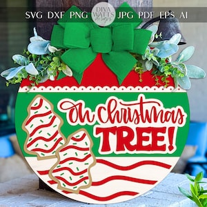 Puede incluir: Adorno navideño redondo con lazo verde y vegetación. El adorno tiene un diseño rojo y verde con las palabras "Oh Christmas TREE!" y dos galletas de jengibre. El adorno es de madera.