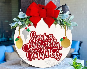 May include: A round wooden Christmas sign with the text "Have a Holly Jolly Christmas" in red script. It features a large red bow, greenery, and gold and silver ornament cutouts. A bell hangs above.