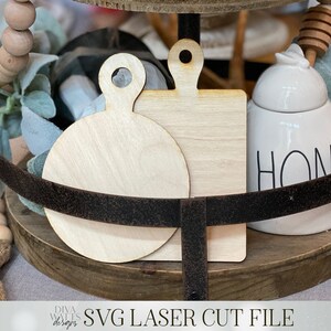 May include: Two unfinished wooden cutting boards, one round and one square, with a handle on each. The boards are on a tiered wooden tray with a white ceramic honey pot with the word "HONEY" on it.