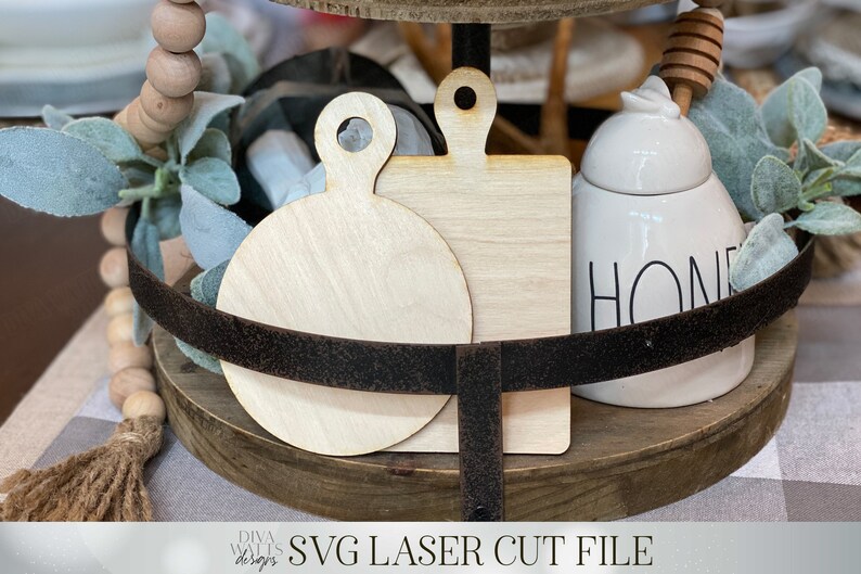 May include: Two unfinished wooden cutting boards, one round and one square, with a handle on each. The boards are on a tiered wooden tray with a white ceramic honey pot with the word "HONEY" on it.