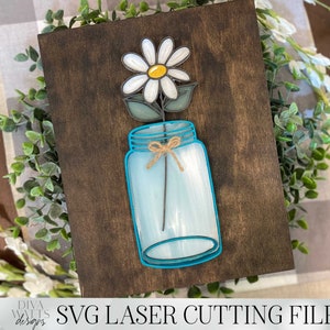 May include: A white daisy with a yellow center is in a blue mason jar with a brown twine bow. The jar is on a brown wooden background.