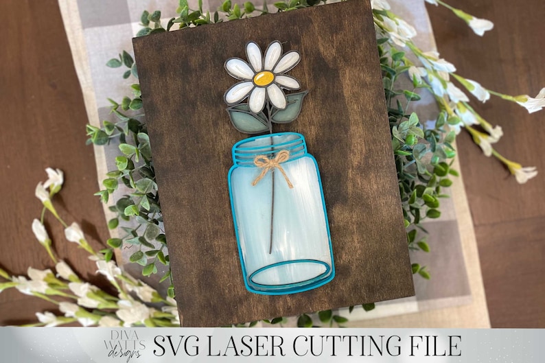 May include: A white daisy with a yellow center is in a blue mason jar with a brown twine bow. The jar is on a brown wooden background.