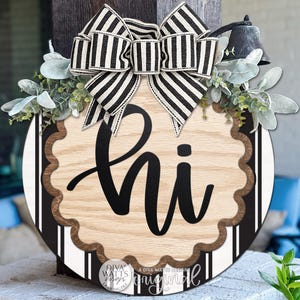 May include: A round wooden door sign featuring a light wood grain center with the black script text "hi" surrounded by a scalloped brown border. The background has black and white vertical stripes, topped with a black and white striped bow.