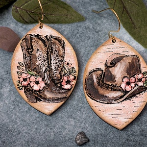 May include: A pair of wooden earrings with a rustic, birch-like finish. One earring features a pair of brown cowboy boots with pink flowers, while the other earring features a brown cowboy hat with pink flowers.