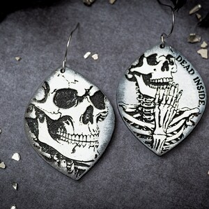 May include: A pair of silver earrings with a black and white skull design. One earring features a skull with a toothy grin, while the other earring features a skeleton with the text "DEAD INSIDE".